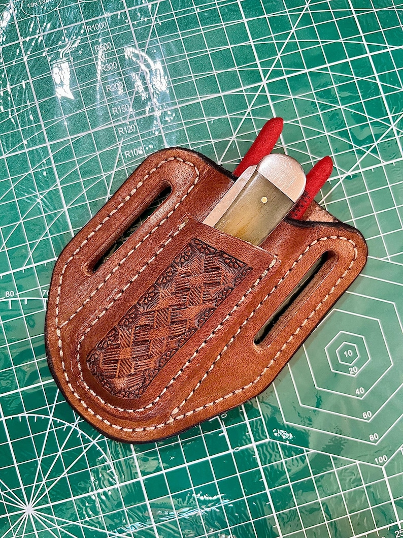 May include: A brown leather knife sheath with a decorative embossed pattern and white stitching. A knife with a light-colored handle and red accents is partially visible. The sheath is on a green cutting mat.