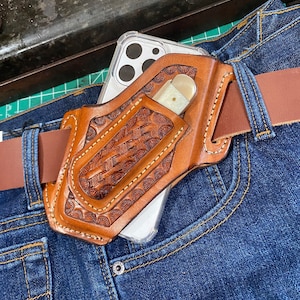 Op de afbeelding: Een bruin leren holster met een decoratief reliëfpatroon, met een smartphone en een klein mes. De holster is bevestigd aan een bruine leren riem, gedragen met een blauwe spijkerbroek.