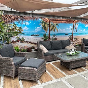 May include: A rattan garden furniture set with dark grey cushions, comprising a sofa, two armchairs, an ottoman, and a glass-topped coffee table. The backdrop is a beach scene mural, with a brown canopy overhead.
