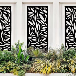 May include: Three black metal privacy screens with a leaf design are mounted on a white wall. The screens are surrounded by lush green plants.