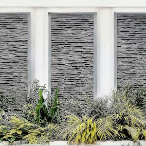 May include: An outdoor wall design with three rectangular sections clad in textured grey stone. White pillars separate the sections. The base is planted with lush green and yellow foliage, providing a natural contrast to the stone.