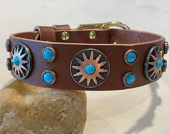 May include: A brown leather collar featuring circular metal sunburst ornaments with embedded turquoise stones. The collar includes smaller round turquoise accents between the sunbursts, brass hardware, and two gold-toned rivets near the buckle.