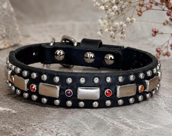 May include: Black leather dog collar with silver-tone studs and rectangular accents. The collar features small, round studs and larger rectangular silver accents, with red, purple, and orange gems. A silver buckle and D-ring are visible.