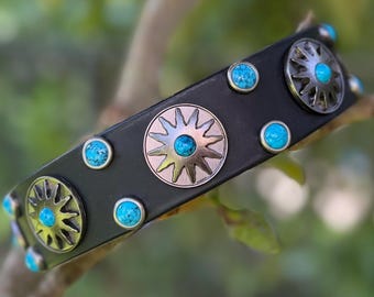 May include: A black leather cuff bracelet featuring silver-toned sunburst metal conchos and small, round, bright blue turquoise cabochons. The design alternates between larger sun-shaped metal accents and smaller circular turquoise stones.
