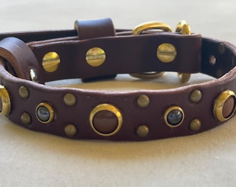 May include: A dark brown leather collar featuring gold-toned metal hardware, including a buckle and D-ring. The strap is embellished with alternating round brown and dark grey cabochon studs set in gold-toned bezels, along with smaller brass rivets.