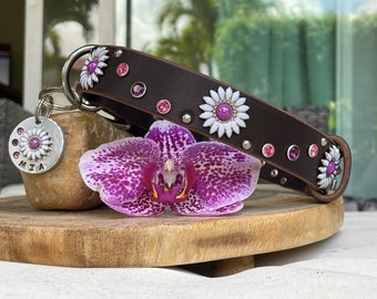 May include: A brown leather dog collar with white daisy accents and pink crystal embellishments. A silver tag with the name "MIA" is attached. A purple and white orchid flower rests near the collar, displayed on a wooden surface.