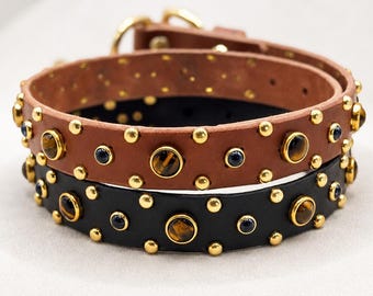 May include: Two leather collars, one tan and one black, each featuring gold-tone metal studs and circular tiger eye stone inlays. The collars are coiled, showing a gold-tone metal buckle and D-ring hardware at the top of the tan leather strap.