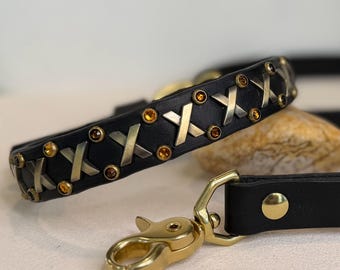 May include: A black leather collar and leash set featuring brass-toned X-shaped metal accents and small amber-colored circular crystals embedded along the length of the collar, finished with a polished brass swivel snap hook and rivets.