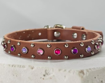 May include: A brown leather collar featuring a series of round, multi-colored translucent gemstones in shades of pink and purple, interspersed with small silver-toned metal studs along the entire length of the band and a metal buckle closure.