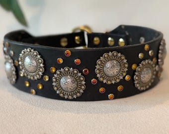 May include: A black leather collar featuring circular silver-toned metal conchos with intricate floral engravings. The collar is embellished with small, round, amber-colored crystals interspersed between the conchos and secured with brass-toned rivets.