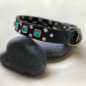 May include: A black leather collar featuring square-cut teal gemstones set in silver-toned metal frames, interspersed with small circular clear crystals. The collar includes a silver-toned metal D-ring and a buckle closure mechanism.