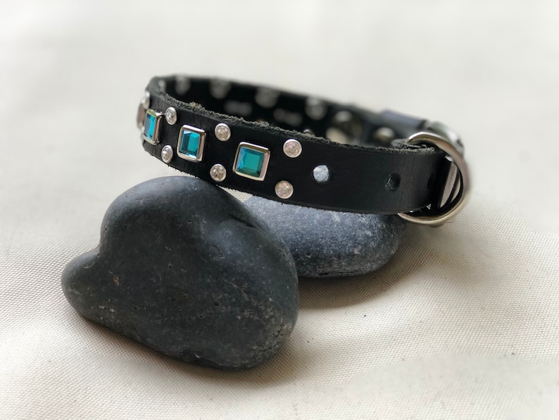 May include: A black leather collar featuring square-cut teal gemstones set in silver-toned metal frames, interspersed with small circular clear crystals. The collar includes a silver-toned metal D-ring and a buckle closure mechanism.