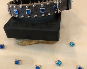 May include: A gray leather dog collar with silver studs and blue square rhinestones. The collar has a silver buckle and is shown on a white surface with several loose rhinestones.