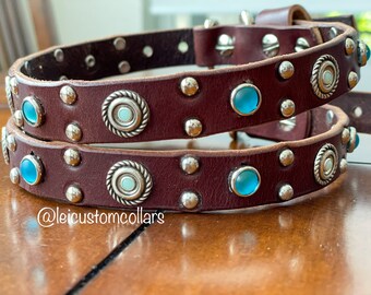 May include: A brown leather dog collar with silver studs and turquoise blue gemstones. The collar is double layered and has a silver buckle. The collar is decorated with silver circles with a white center.