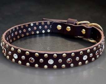 May include: A dark brown leather collar featuring a series of alternating metallic studs and circular crystals in clear and amber tones. The collar includes a gold-toned metal buckle, a matching D-ring, and a leather keeper loop for secure fastening.