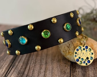 May include: A black leather collar featuring gold-tone metal studs and embedded green and blue circular gemstones. A round gold-tone metal tag is attached to the collar, engraved with the word BONES and decorated with small blue and green crystals.