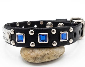 May include: Black leather dog collar with silver studs and three square blue gemstones. The collar has a metal buckle and a small metal tag with engraved text. The collar is resting on a small, light-colored stone.