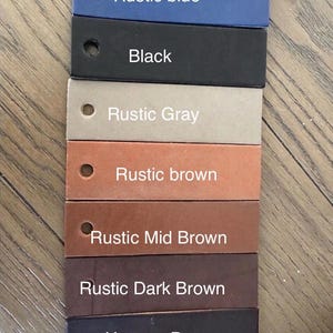 May include: A set of seven rectangular leather swatches arranged in a vertical column, each featuring a circular hole on the left side. The colors are labeled as Rustic blue, Black, Rustic Gray, Rustic brown, Rustic Mid Brown, Rustic Dark Brown, and Havana Brown.
