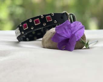 May include: A black leather dog collar with silver studs and pink rhinestones. The collar is lying on a white surface next to a purple flower and a gray rock.