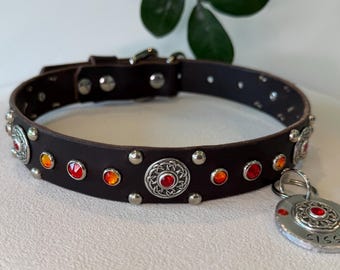 May include: A dark brown leather collar featuring silver-toned metal studs, circular decorative plates with intricate knotwork patterns, and embedded orange and red faceted crystals. A matching circular metal tag is attached, engraved with SISSY.