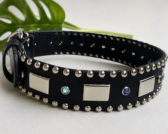 May include: A black leather collar featuring two rows of silver-toned metal studs along the edges, alternating rectangular silver plates, and small circular blue and green gemstones embedded between the plates, with a silver metal buckle.