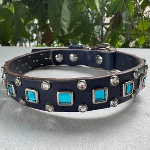 May include: A dark blue leather collar featuring a series of square-cut turquoise gemstones and round clear crystals set in silver-toned metal bezels. The collar includes a silver-toned metal buckle and rivets, providing a secure closure mechanism.