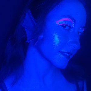 Könnte beinhalten: Eine Person mit spitzen Ohrprothesen und neonpinkem grafischem Eyeliner unter blauem UV-Licht. Das Make-up zeichnet sich durch scharfe, winklige Linien um die Augen aus, und die Person trägt ein schwarzes Kleidungsstück mit Riemen vor dunklem Hintergrund.