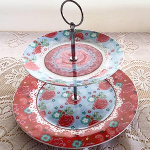 May include: A two-tiered serving stand featuring two round ceramic plates with a floral pattern in shades of red, light blue, and white. The plates are connected by a central silver-coloured metal rod with a circular handle at the top.