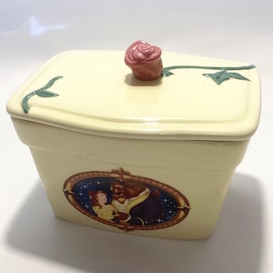 May include: A cream ceramic box with a lid decorated with a pink rose and green leaves. The box features an oval image of Belle and the Beast. A decorative storage item.