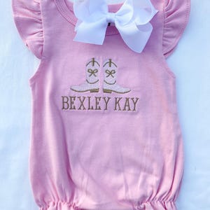 May include: A pale pink baby romper with frilled sleeves and a gathered hem. The romper has an embroidered design of cowboy boots and the text "BEXLEY KAY" in brown thread. A large white bow is attached at the neckline.