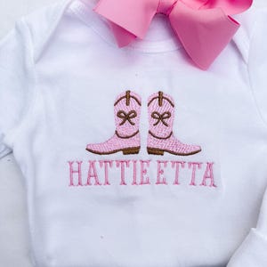 May include: White baby bodysuit featuring embroidered pink cowboy boots and the name "HATTIE ETTA" in pink. A large pink bow is attached. The garment is designed for infants.