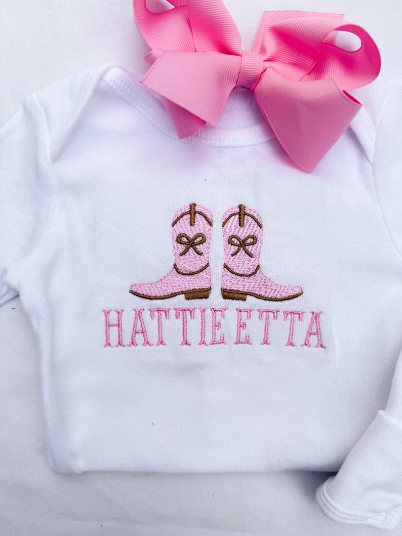 May include: White baby bodysuit featuring embroidered pink cowboy boots and the name "HATTIE ETTA" in pink. A large pink bow is attached. The garment is designed for infants.