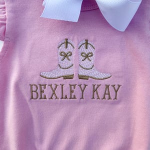 May include: Pale pink baby outfit featuring embroidered cowboy boots and the name "BEXLEY KAY" in brown thread. A large white bow is attached. The outfit is made of soft material.