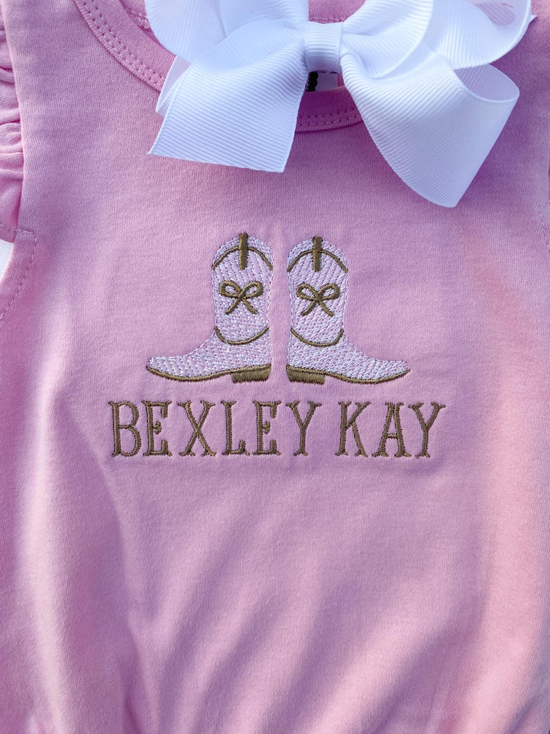 May include: Pale pink baby outfit featuring embroidered cowboy boots and the name "BEXLEY KAY" in brown thread. A large white bow is attached. The outfit is made of soft material.