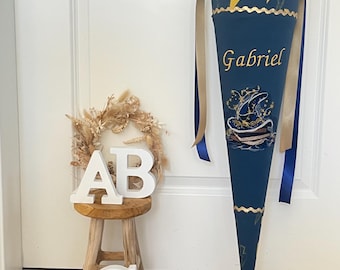 May include: A dark blue fabric cone with a yellow lightning bolt pattern, tied with gold and blue ribbons. The name Gabriel is embroidered in yellow, featuring a wizard hat graphic. Beside it, a small wooden stool holds white letters A, B, and C.