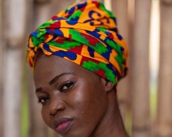 May include: A woman with dark skin is wearing a colorful head wrap with a yellow, green, blue, and red pattern. She is looking at the camera with a serious expression. She is wearing a white shirt.