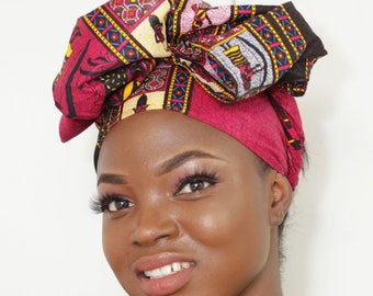 May include: A woman with dark skin is wearing a red, black, and yellow patterned head wrap. The wrap is tied in a large bow on top of her head. She is smiling and looking at the camera.