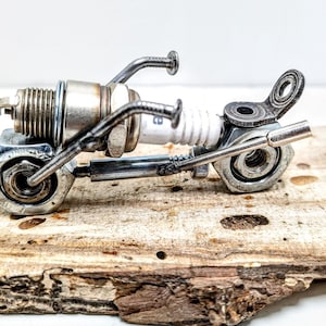 May include: A miniature motorcycle sculpture made from recycled metal components, including spark plugs, nuts, and bolts. The silver and grey parts are welded together to create the bike's frame, wheels, and handlebars. The sculpture is mounted on a piece of natural wood.