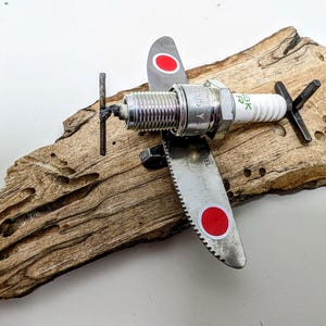 May include: A handcrafted airplane sculpture made from a spark plug, metal saw blades, and other metal components. The wings feature red circles. The sculpture is mounted on a piece of weathered wood, creating a unique industrial art piece.