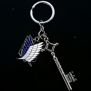 Puede incluir: Un llavero plateado con una llave y un emblema alado. La llave tiene detalles intrincados, y el emblema muestra alas blancas con acentos azules. El llavero está suspendido de un anillo plateado, sobre un fondo negro.