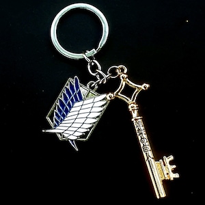 May include: A silver keychain with two charms. One is a gold-coloured key with Japanese text. The other is a rectangular charm with a white wing design and blue accents. The keychain is on a black background.