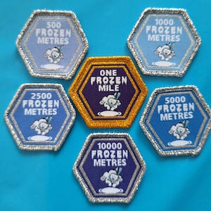 May include: A set of embroidered hexagon patches with silver edging. The patches display distances in metres and miles, including "500 Frozen Metres" and "One Frozen Mile." Each patch features a cartoon snowman illustration.