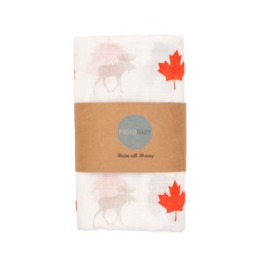May include: White muslin baby swaddle blanket featuring a red maple leaf and moose design. The blanket has the words "Proud Baby" and "Made with Meaning" printed on a brown label.
