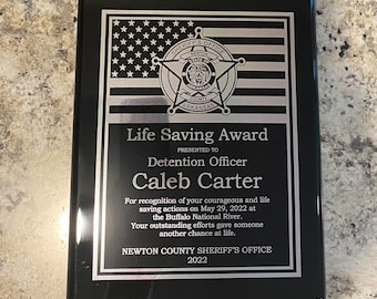 May include: A black plaque with a silver border and a silver star with the words "Sheriff's Office" and "Newton County Arkansas" on it. The plaque reads "Life Saving Award" and "Presented to Detention Officer Caleb Carter" and includes a description of the award.