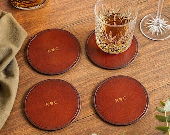 May include: Four circular, brown leather coasters with a lighter brown border and the initials "R ♡ C" in gold. A glass of amber liquid sits on one coaster. The coasters are on a wooden surface.