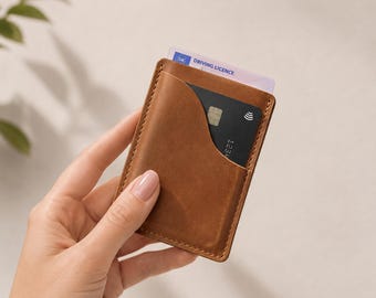 May include: A tan leather card holder with visible stitching along the edges. The card holder features a curved pocket design and contains a black payment card and a UK driving licence card partially inserted into the top slot.