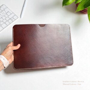 May include: A brown leather laptop sleeve with tan stitching. The rectangular case has a textured surface and a small cutout at the top. The sleeve is held by a person's hand. The text on the image says "Leather Colour: Brown" and "Thread Colour: Tan".