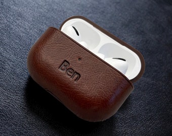 May include: A brown leather AirPod case with the name "Ben" embossed on the front. The case is open, showing white AirPods inside. The case has a textured appearance and a dark brown interior.