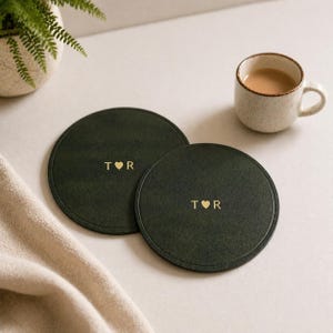 May include: Two dark green circular coasters with gold lettering and a heart symbol. Each coaster has the letters "T R". A small ceramic mug with a brown liquid sits nearby. A beige cloth and a potted plant are in the background.