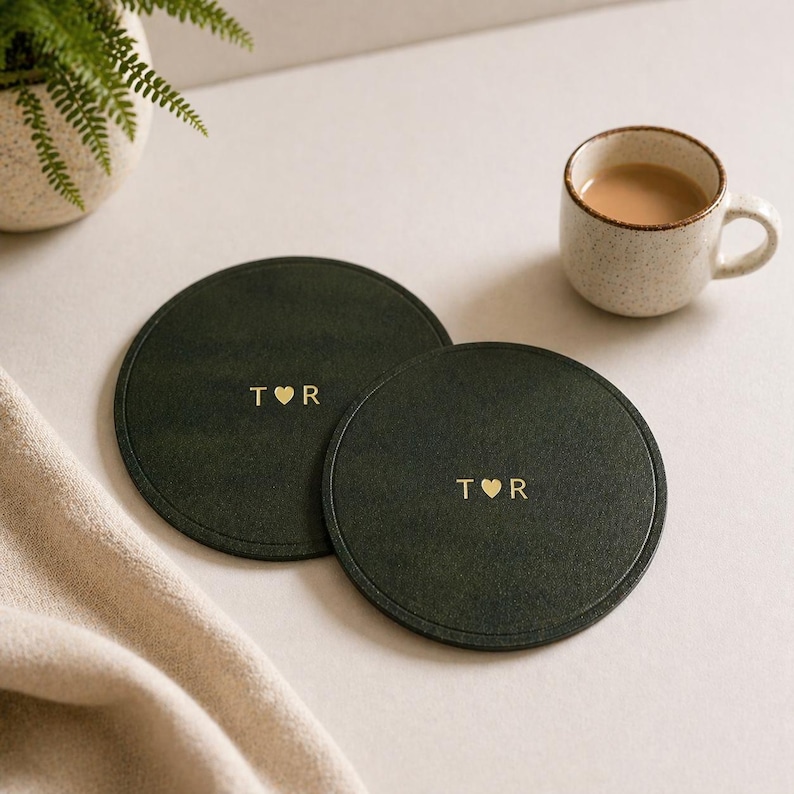 May include: Two dark green circular coasters with gold lettering and a heart symbol. Each coaster has the letters "T R". A small ceramic mug with a brown liquid sits nearby. A beige cloth and a potted plant are in the background.